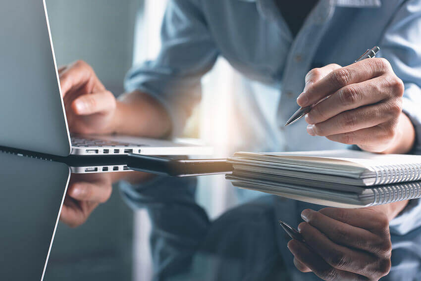 Cropped photo of middle aged Caucasian woman's torso, arms and hands seated in a blue button down shirt at a mirrored desk with a laptop, phone and notepad. Pen in one hand and the other hand on the keyboard. 